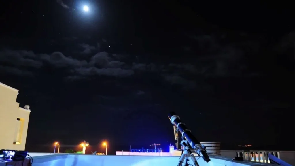 Une éclipse, mille façons de la vivre avec Barceló Hotel Group Télescope installé sur un toit, pointé vers une Lune brillante dans un ciel étoilé parsemé de nuages, évoquant une séance d’observation astronomique nocturne avec lampadaires et balustrade en arrière-plan.