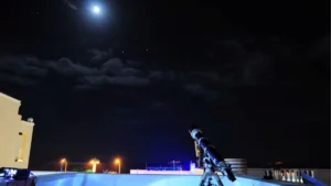 Télescope installé sur un toit, pointé vers une Lune brillante dans un ciel étoilé parsemé de nuages, évoquant une séance d’observation astronomique nocturne avec lampadaires et balustrade en arrière-plan.