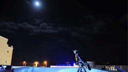Télescope installé sur un toit, pointé vers une Lune brillante dans un ciel étoilé parsemé de nuages, évoquant une séance d’observation astronomique nocturne avec lampadaires et balustrade en arrière-plan.