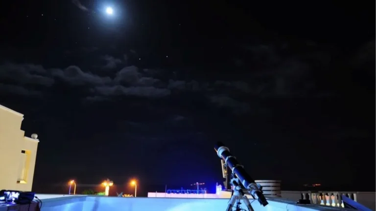 Une éclipse, mille façons de la vivre avec Barceló Hotel Group Télescope installé sur un toit, pointé vers une Lune brillante dans un ciel étoilé parsemé de nuages, évoquant une séance d’observation astronomique nocturne avec lampadaires et balustrade en arrière-plan.