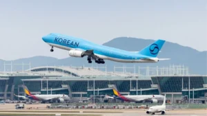 Avion de Korean Air bleu en plein décollage depuis un terminal d'aéroport international, trains d'atterrissage en rentrée, autres appareils au sol et collines en arrière-plan.