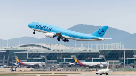 Avion de Korean Air bleu en plein décollage depuis un terminal d'aéroport international, trains d'atterrissage en rentrée, autres appareils au sol et collines en arrière-plan.